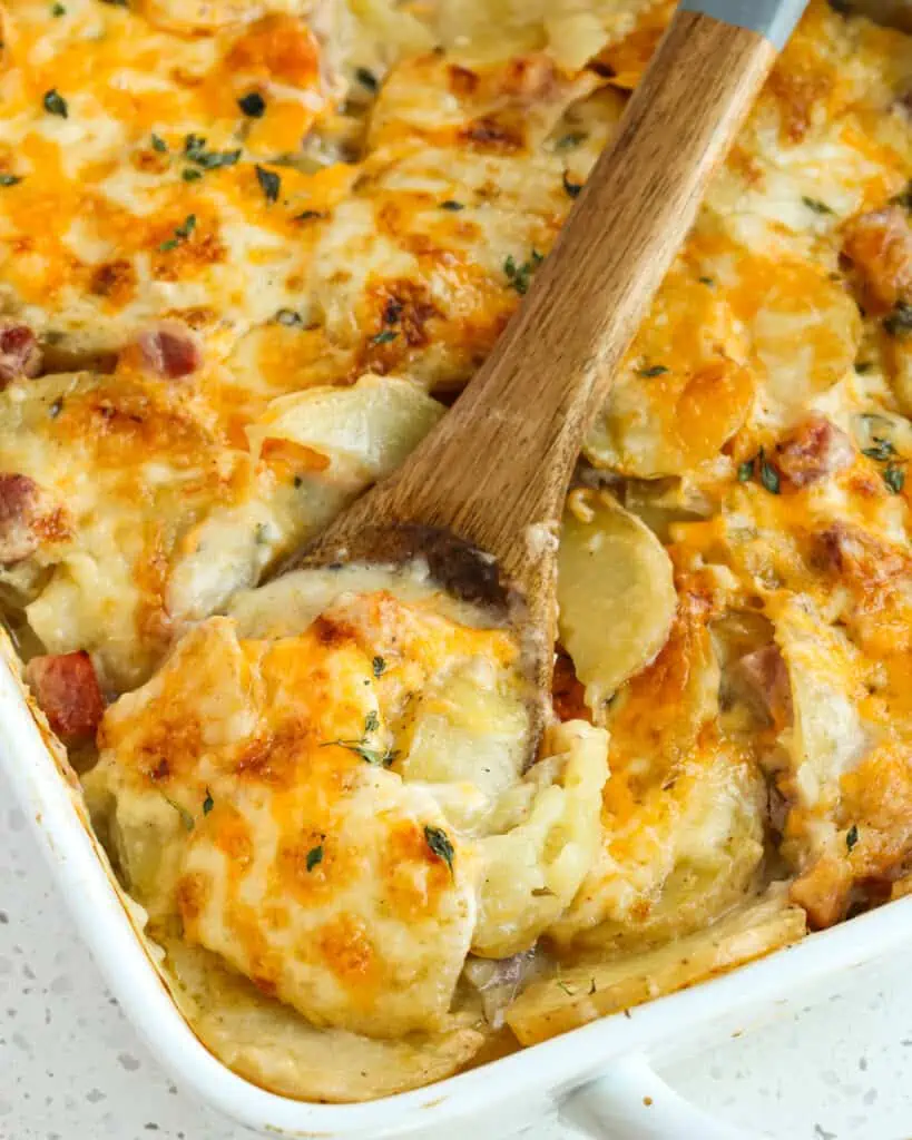 A 9x13 unch casserole dish and and a wooden spoon full of scalloped potatoes and ham. 