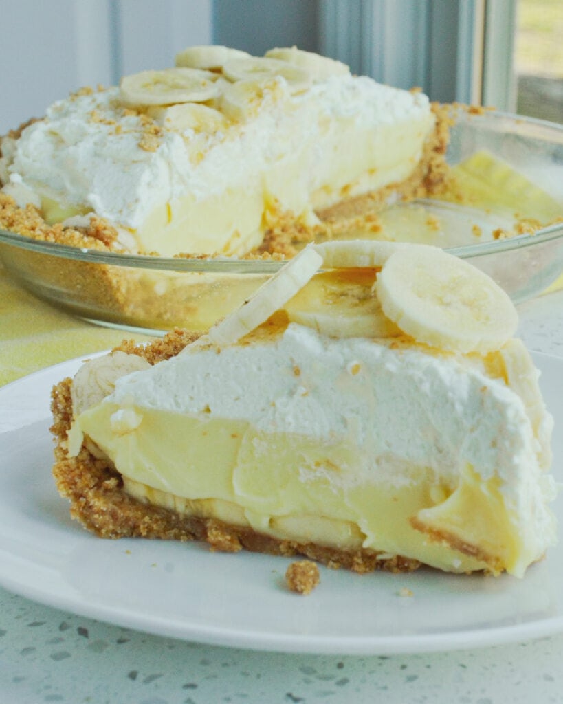 A piece of banana cream pie on a plate with the rest of the pie in the background. 