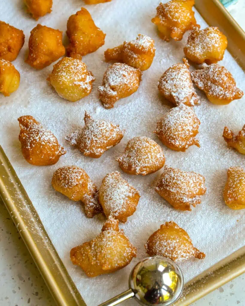 Fried zeppoles sprinkled wiht powdered sugar on a paper towel lined baking sheet. 