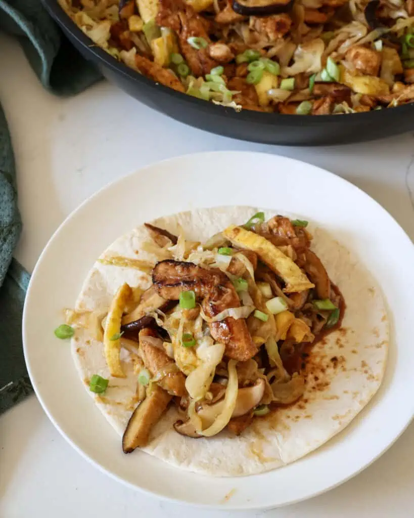 Moo shu chicken on a soft tortilla on a round white single serving plate. 