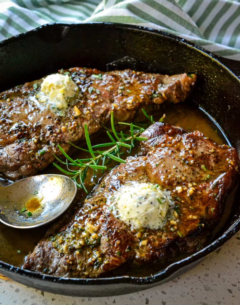 Two New York strip steaks topped with herb buttr in a cast iron skillet. 