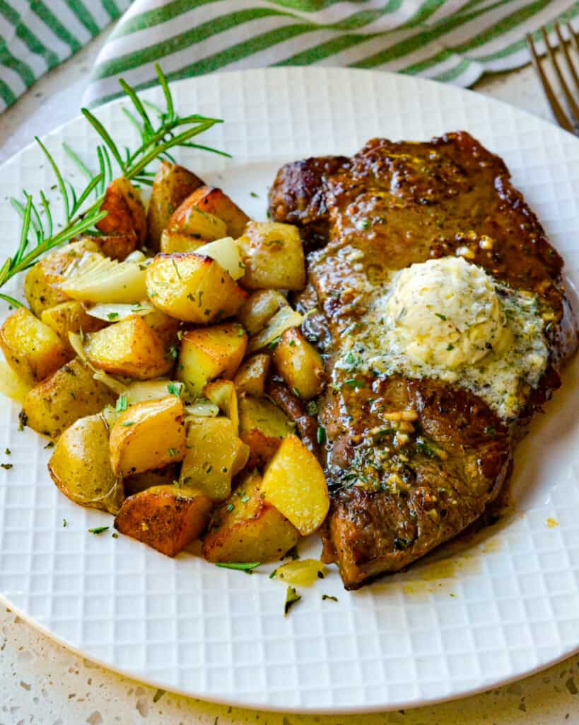 New York strip steak topped with herb butter on a plate with fried potatoes. 