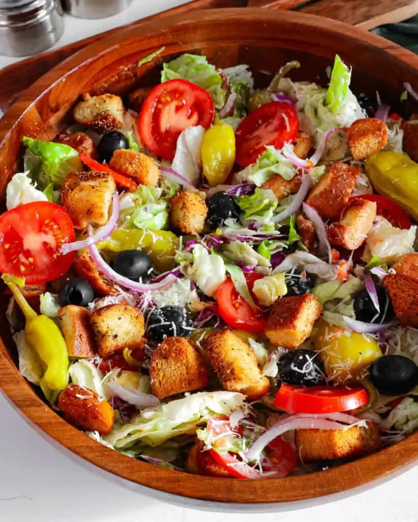 An overhead view of olive garden salad in a wooden bowl with wooden tongs and a salt and pepper shaker in the background. 