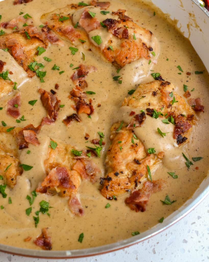 A large skillet full of smothred chicken with crisp bacon, lightly breaded chicken breasts, and lusciously creamy seasoned gravy made from common pantry spices. 