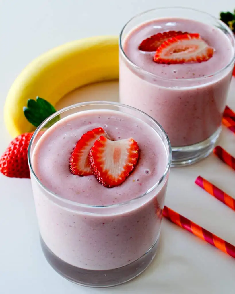 Two glasses full of strawberry banana smoothie with a banana and strawberries in the background.