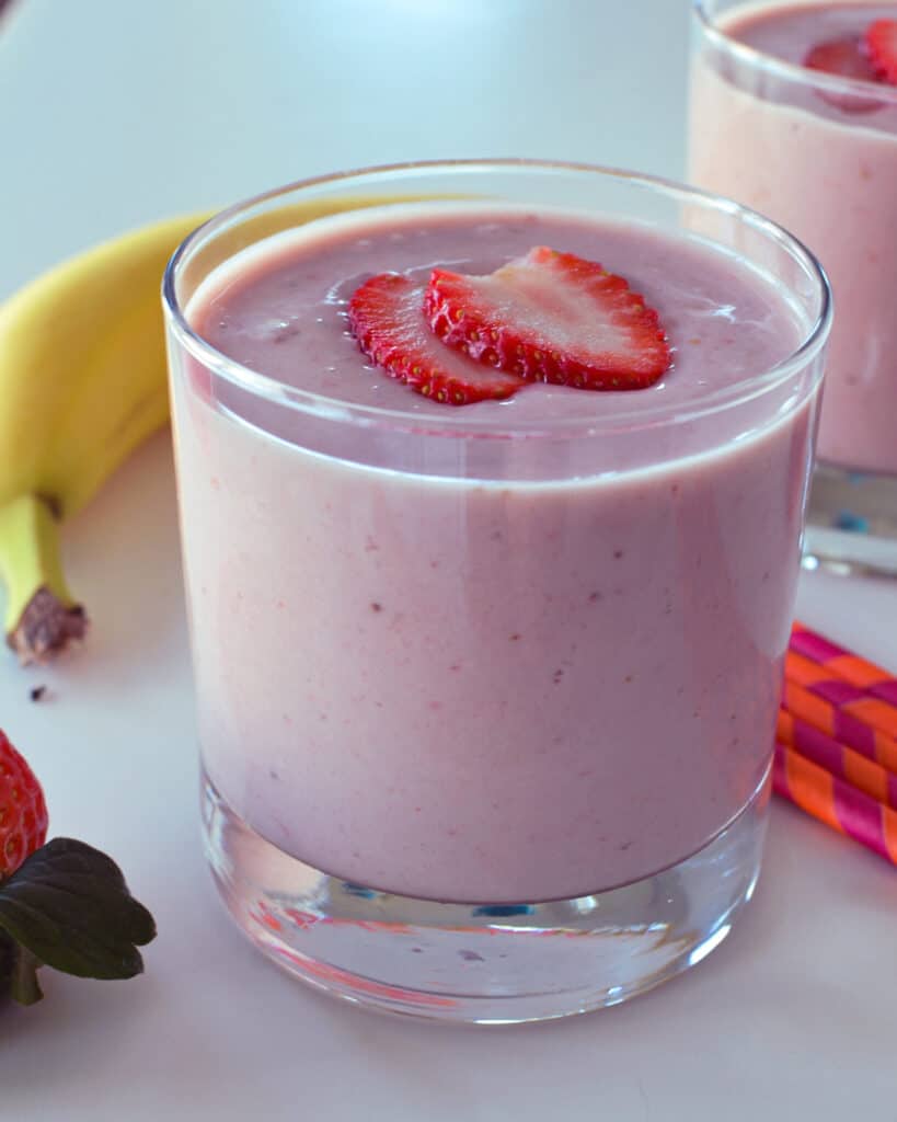 Two strawberry banana smoothies in a glass with a banana and straws in the background. 