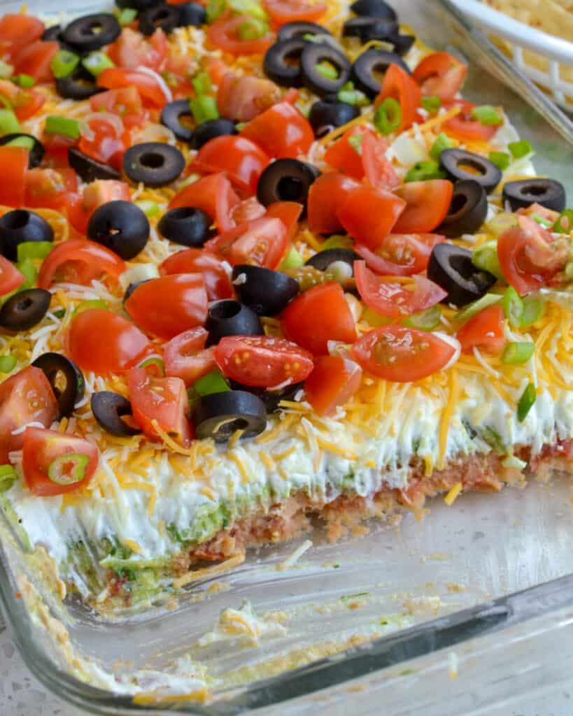 Classic seven layer dip in a glass casserole dish with part of the dip removed so you can see the layers. 