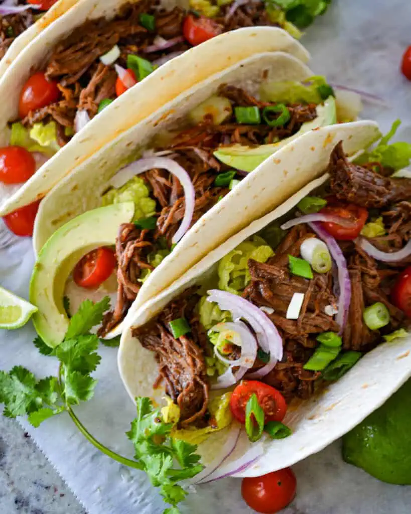 Serve your Beef Barbacoa Tacos in flour tortillas topped with onions, tomatoes, and lettuce