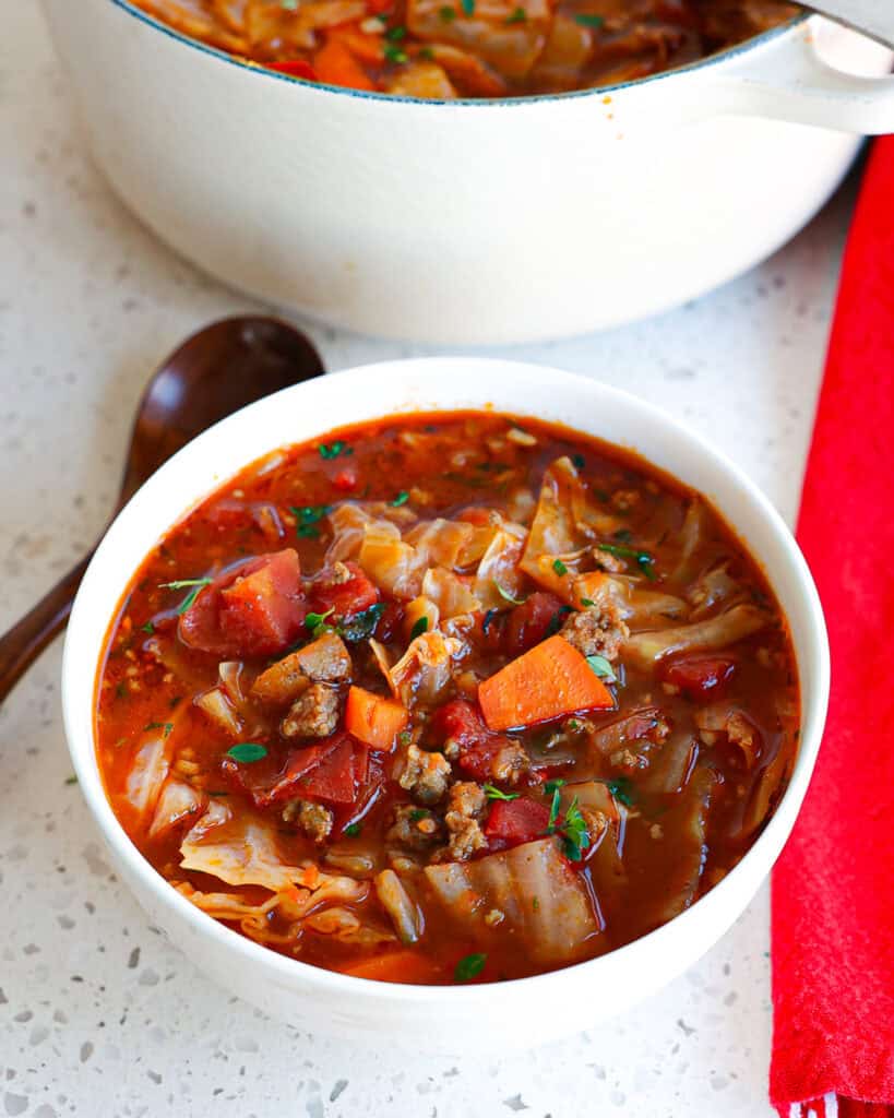 A single serving bowl of cabbage soup with a wooden spoon off to the side and a Dutch oven full of the soup in the background.  
