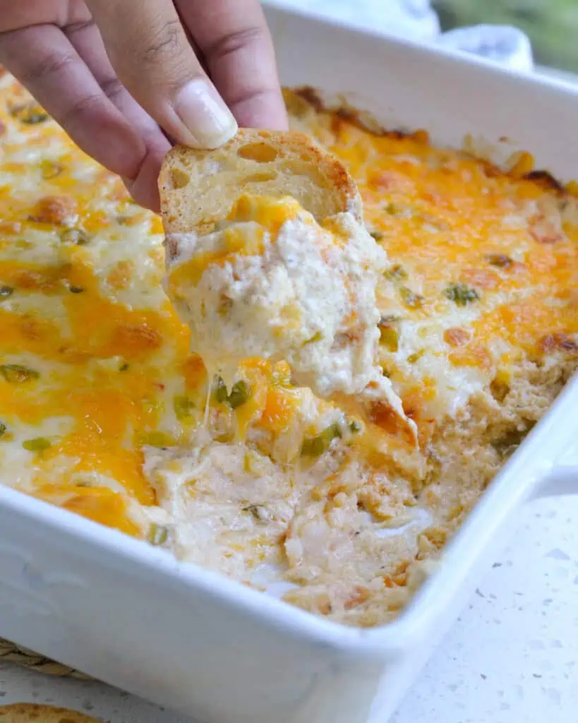 A crostini and casserole dish full of hot crab dip with some of the dip gone and a spreading knife in the dip. 