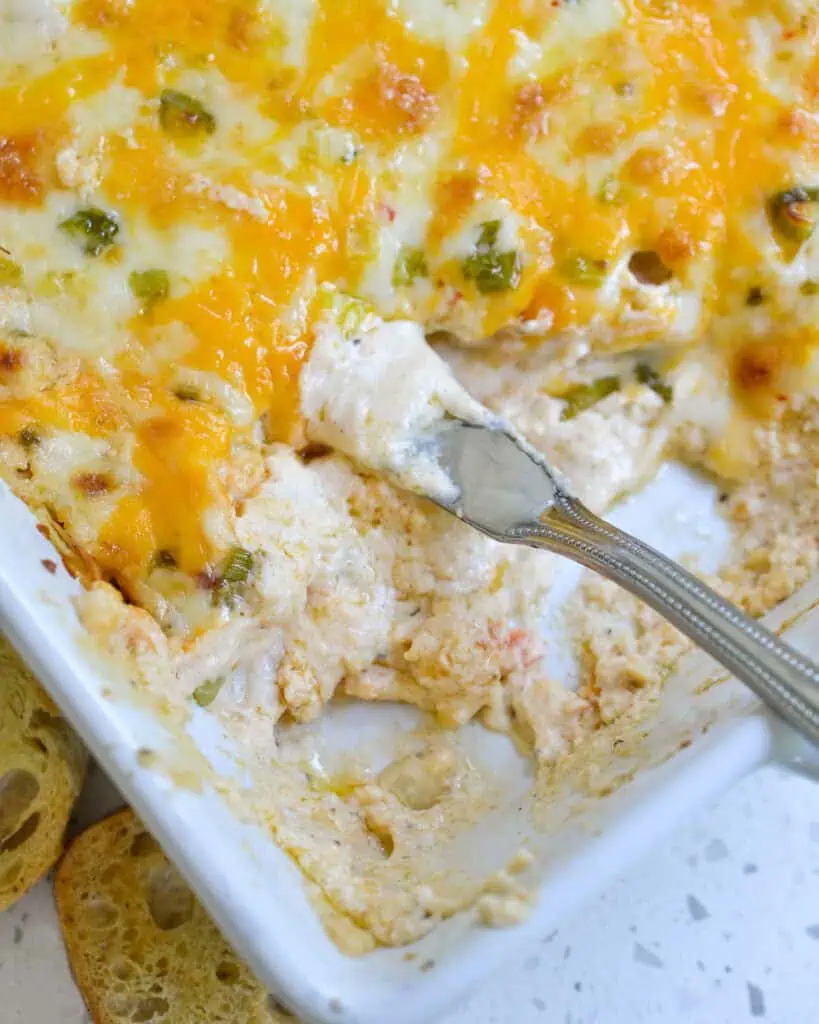 A white casserole dish full of cheese crab dip with some of the dip missing and spreading knife in the dip. 