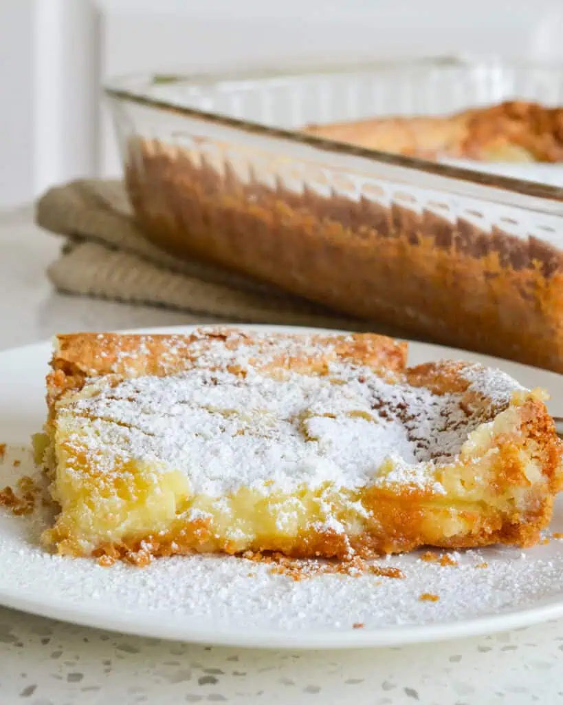 A piece of gooey butter cake sprinkled with powdered sugar on a serving plate with the cake dish in the background. 