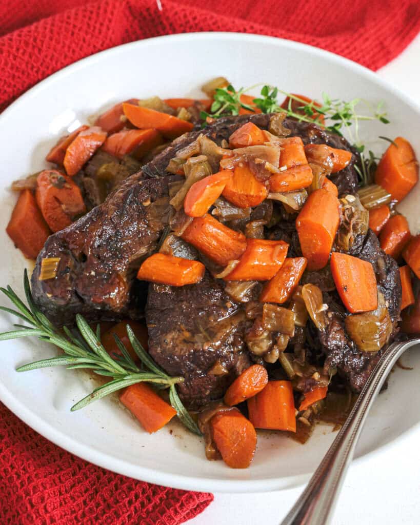 Braised beef, carrots, amd onions in a serving bowl with fresh rosemary and thyme. 