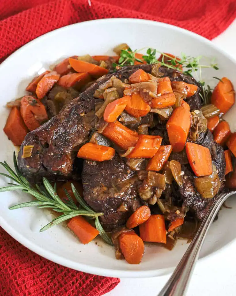 Braised beef, carrots, amd onions in a serving bowl with fresh rosemary and thyme.