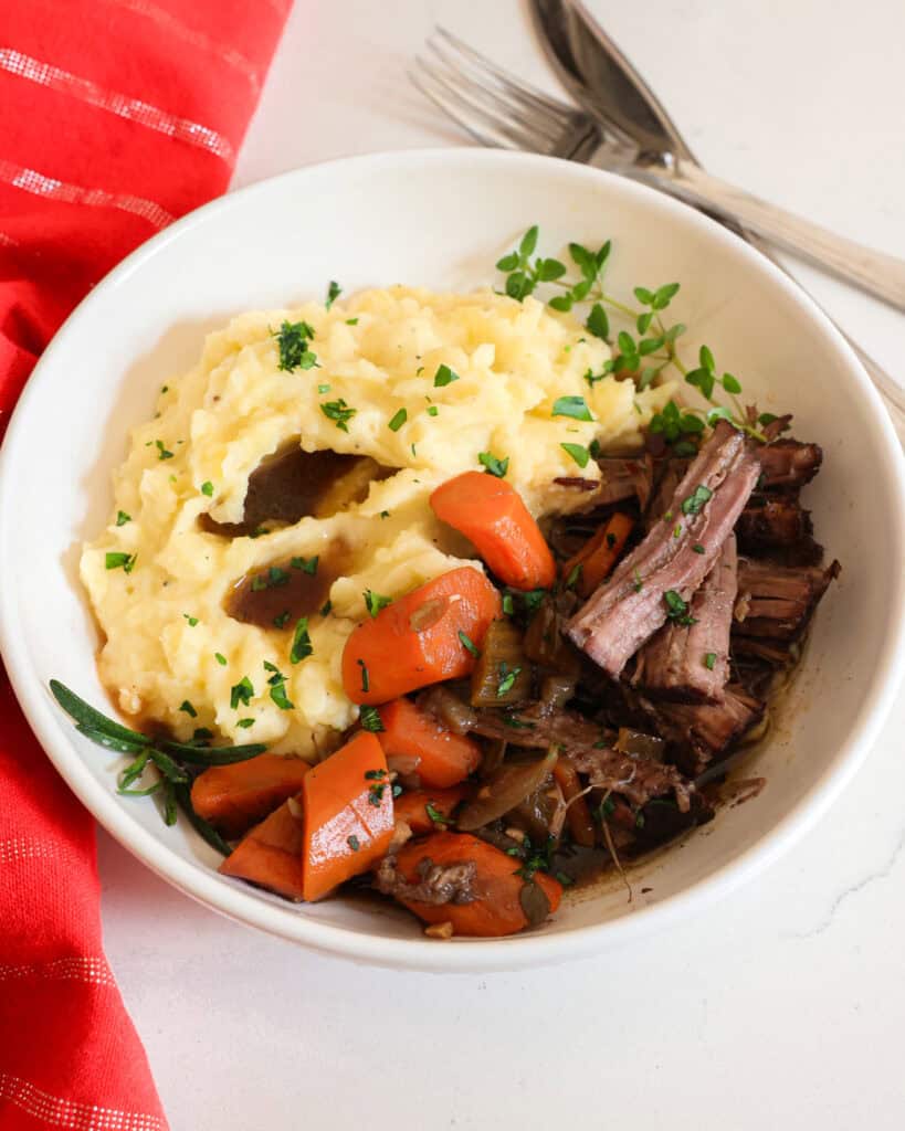 Braised Beef Tender braised beef in a bowl with carrots, mashed potatoes, herbs, and red wine sauce.