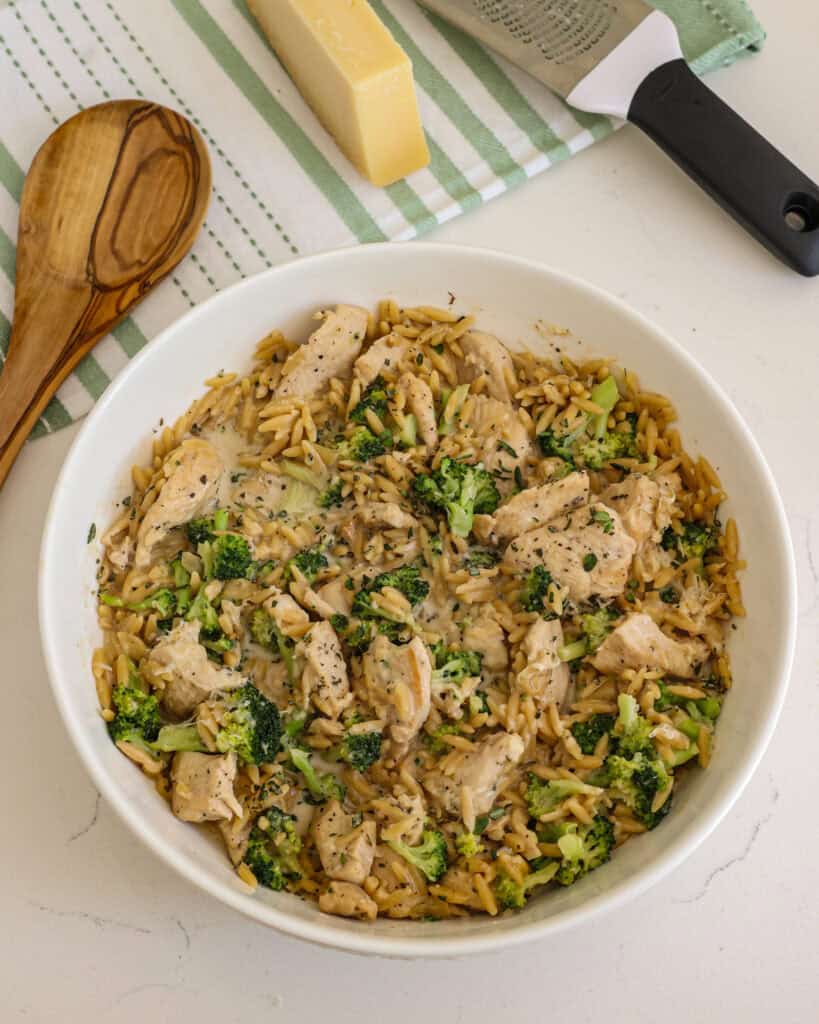 Creamy chicken and orzo in a bowl with a spoon, parmesan cheeese, and cheese grater. 