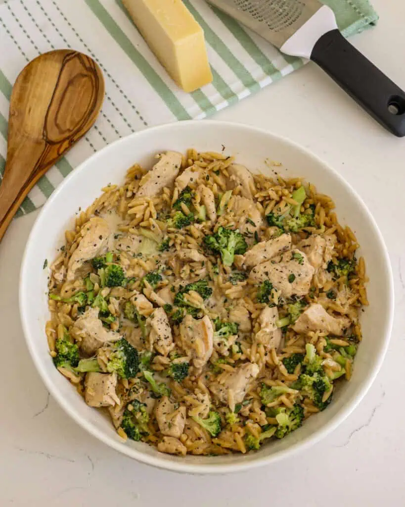 Creamy chicken and orzo in a bowl with a spoon, parmesan cheeese, and cheese grater. 