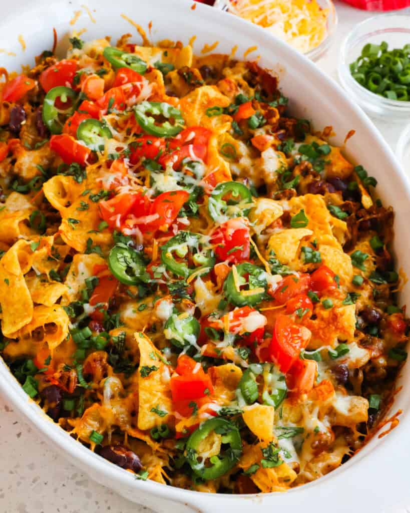 Baked Frito pie in a casserole dish with a small bowl of green onions and shredded cheddar and Montery Jack in the background. 