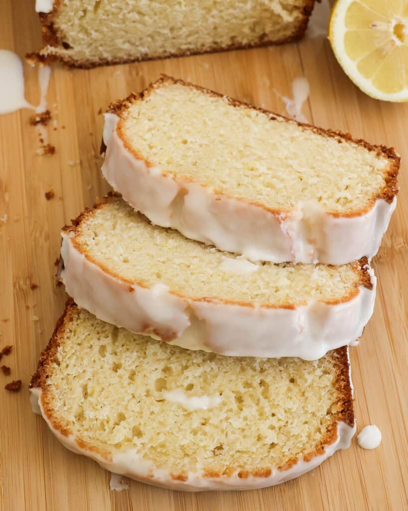 Three slices of lemon bread with glaze on a cutting board with fresh lemons. 