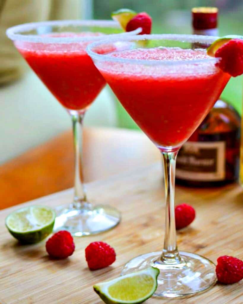 A glass of frozen raspberry margarita garnished with a lime slice and a raspberry in front of a bottle of Grand Marnier.