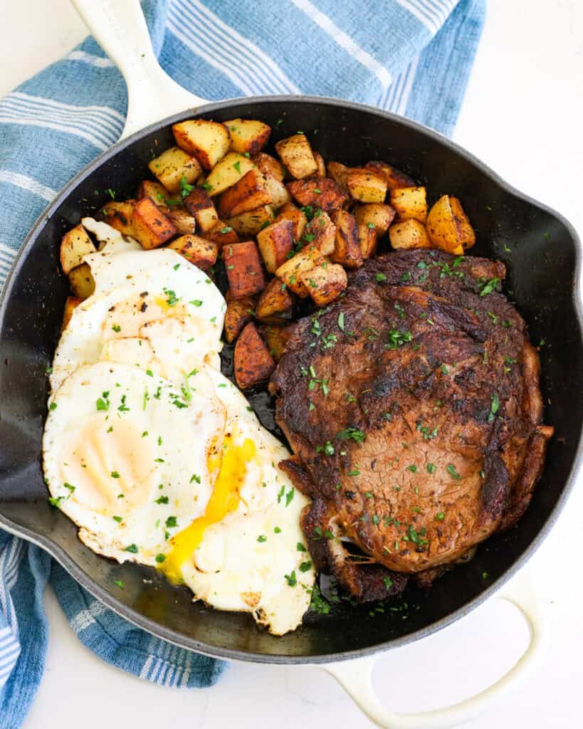 Seasoned steak, fried potatoes, and eggs over easy in a large skillet.