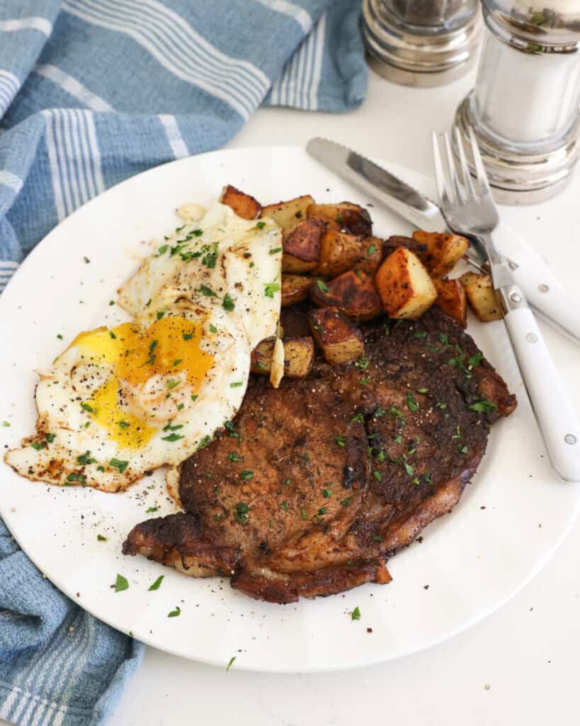 Seasoned seared steak, fried potatoes, and eggs on a plate. with a fork and knfie.