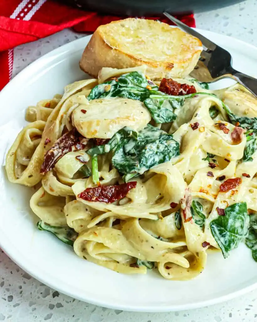 A individual plate full of Tuscan chicken pasta, a fork and a pice of toasted baguette. 