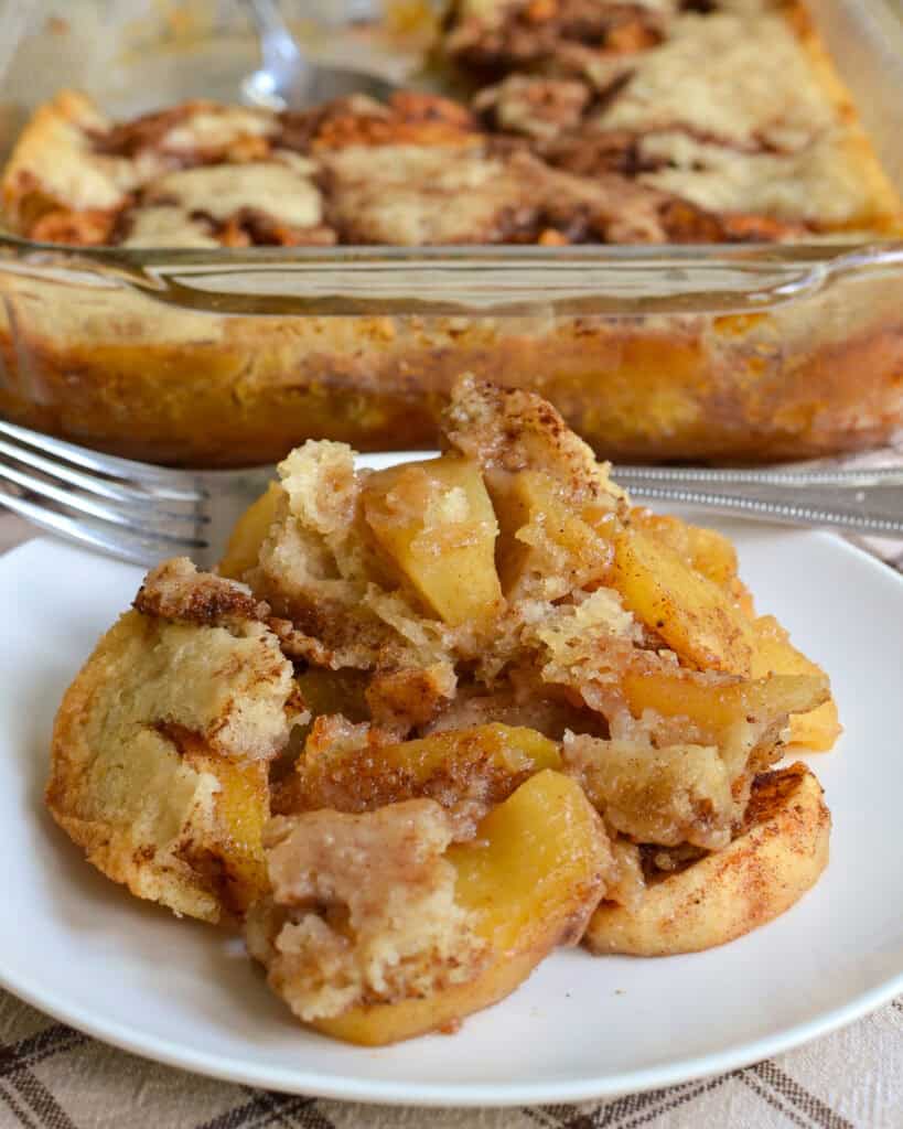 Easy Apple Cobbler Baked apple cobbler on a round plate with a fork and the casserole apple cobbler in the background.