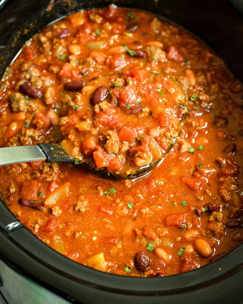 A crock pot and a ladle full of chili with beans and tomatoes.