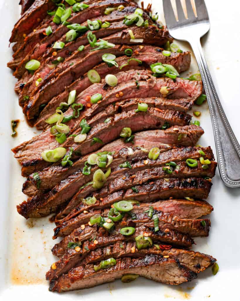 Sliced flank steak on a white serving platter with a serving fork. 