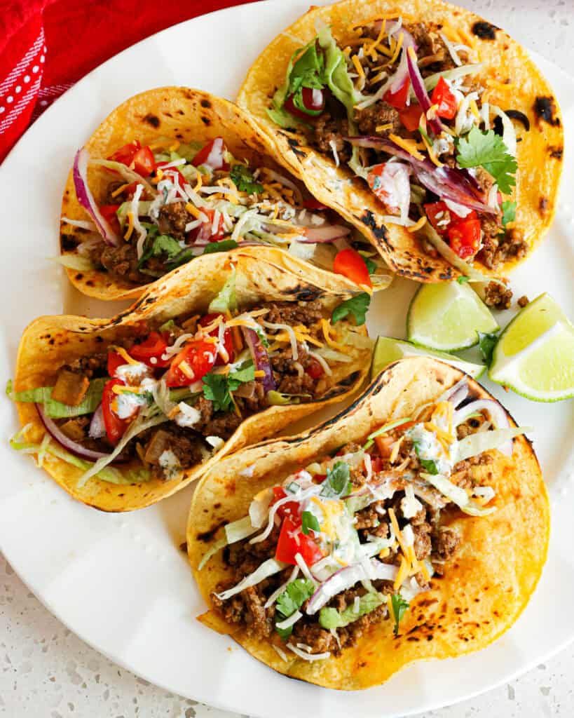Four ground beef tacos on a plate with lettuce. tomato, red onions. and cheddar cheese