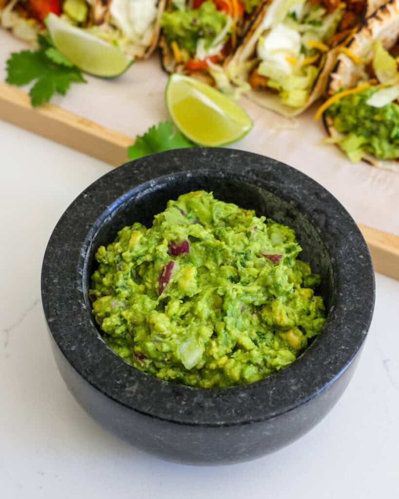 A mortar bowl full of guacamole in front of cutting board full of tacos.