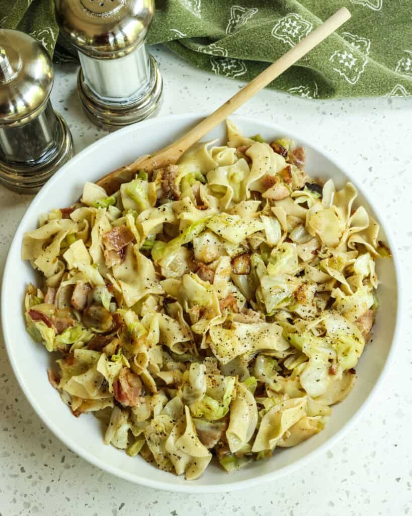 An overhead shot of cabbage and noodles in a bowl with a wooden spoon and salt and pepper shaer in the background.