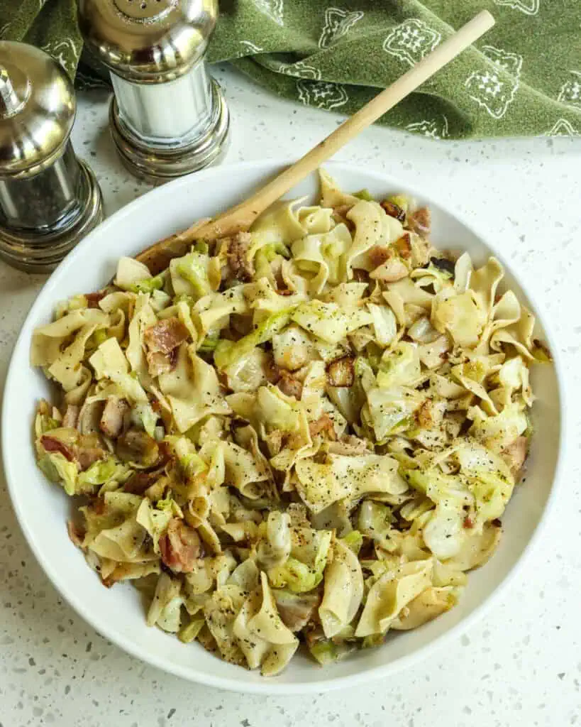 An overhead shot of cabbage and noodles in a bowl with a wooden spoon and salt and pepper shaer in the background. 