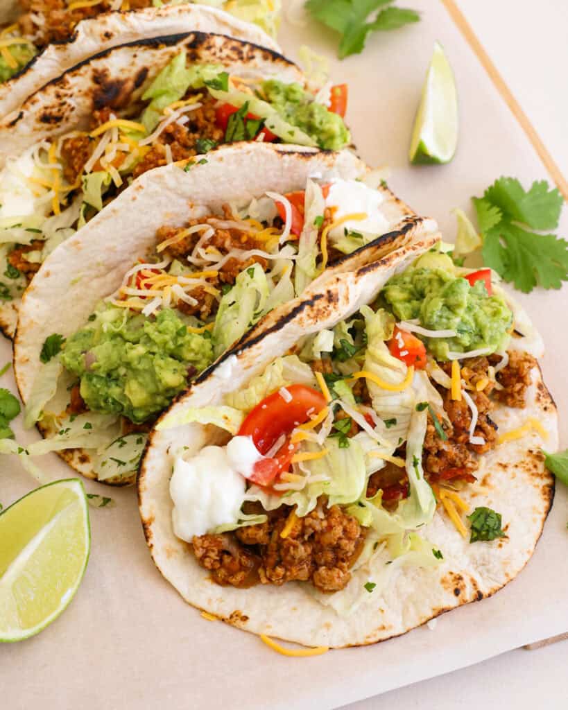 A wooden cutting board full of tacos topped with lettuce, tomatoes, shredded cheese, sour cream, and guacamole. 