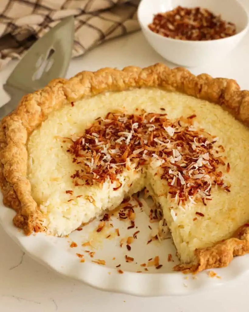 Eine ganze Kokosnuss-Puddingtorte, von der ein Stück entfernt wurde. Im Hintergrund ein Tortenheber und eine Schüssel Kokosnuss.