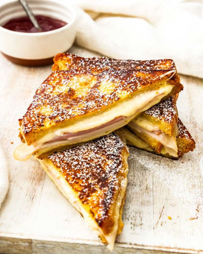 Two halved monte cristo sandwiches on a cutting board with a bowl of raspberry jam in the background.