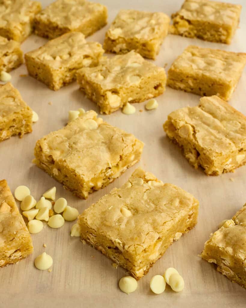 Cut Blondies scattered on a cutting board with white chocolate chips scattered around.