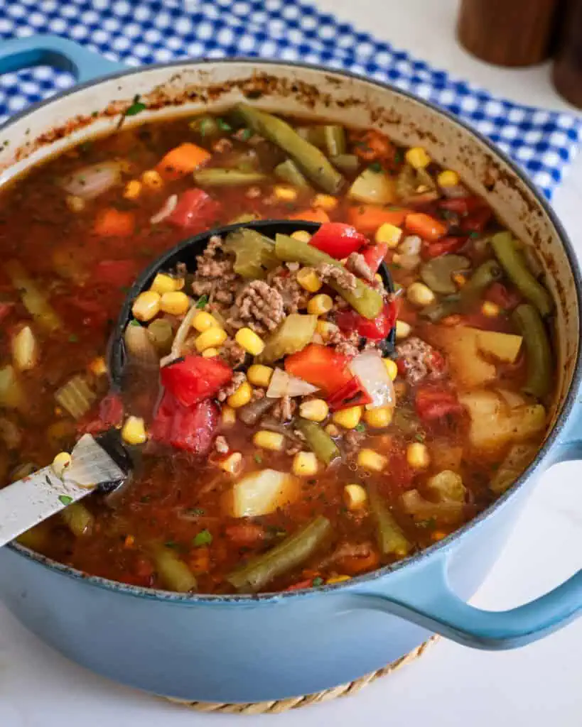 A large soup ladld full of hamburger soup  over a Dutch oven full of hamburger sup. 