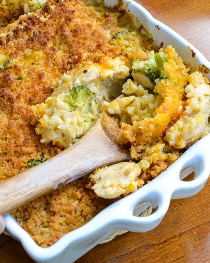 A white 9x13 inch casserole and a wooden spoon filled with chicken broccoli rice cassrole. 
