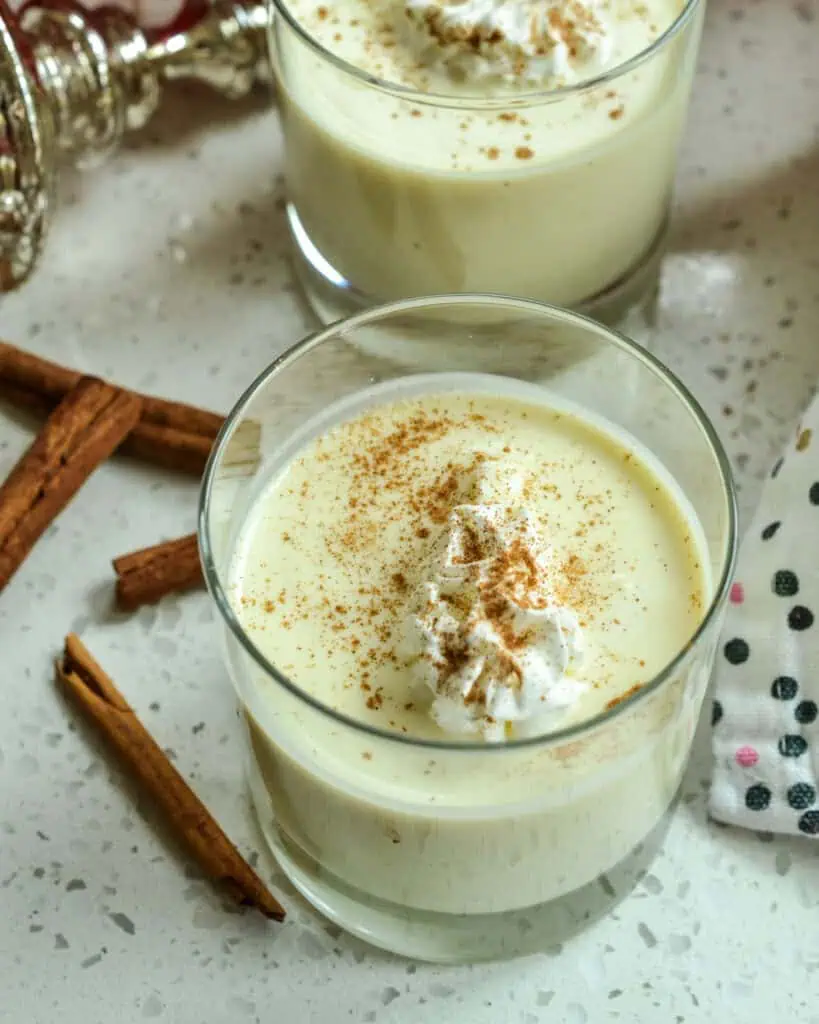 An overhead views of two glasses of eggnog with whipped cream and ground nutmeg. Cinnamon sticks are on the side and an ornament is in the background.