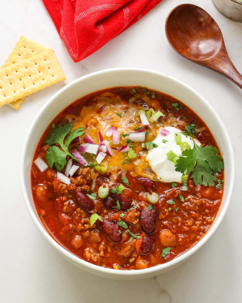 A bowl full of instant pot chili with crackers, a red kitchen towel, and a wooden spoon in the background.
