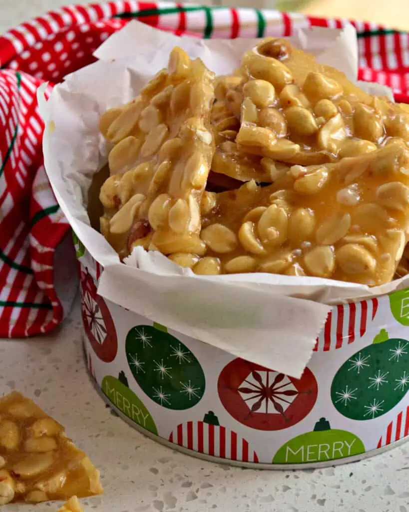 A Christmas tin full of peanut brittle with a red, white, and green towel in the background.