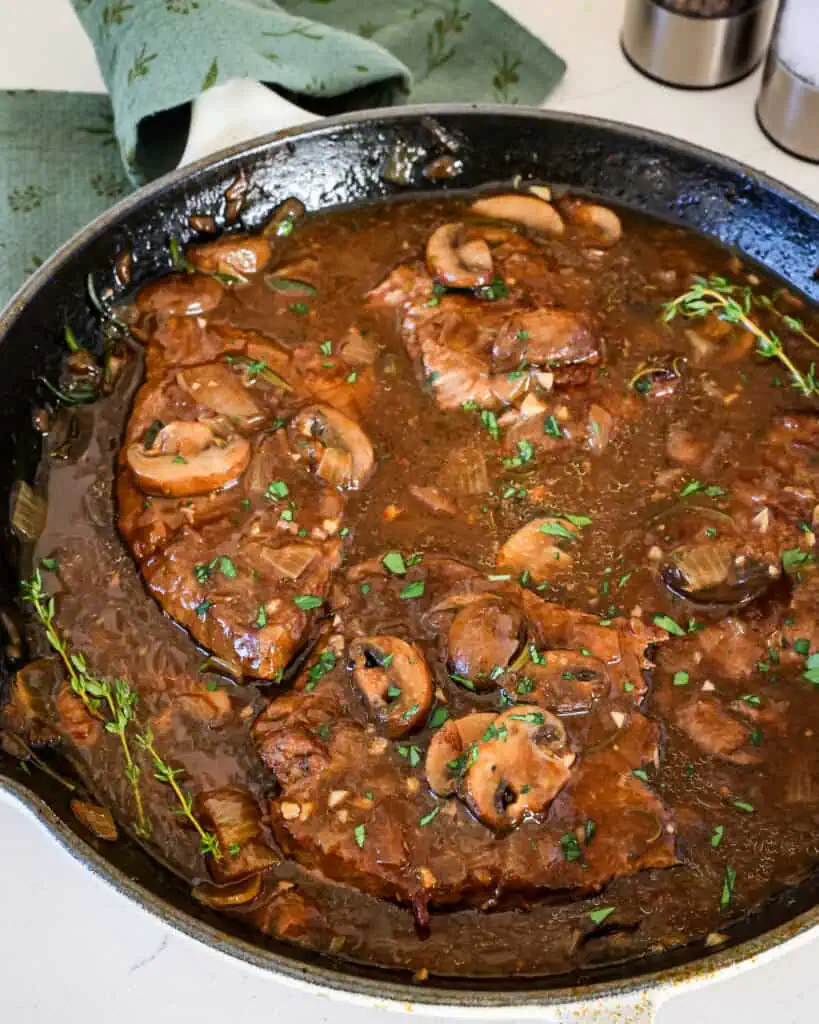 A large cast iron skillet full of round steak and gravy with mushrooms, onions,, and fresh herbs.