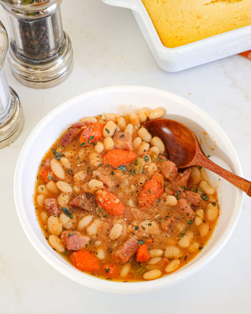 A single serving bowl of ham an beans with a salt and pepper shaker and cornbread in the background.