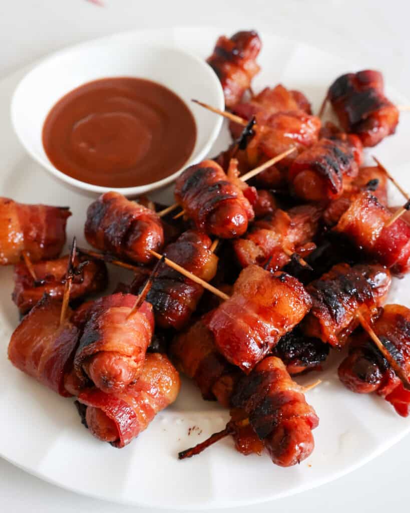 A white plate full of bacon wrapped little smokies basted with brown sugar next to a bowl of BBQ sauce. 