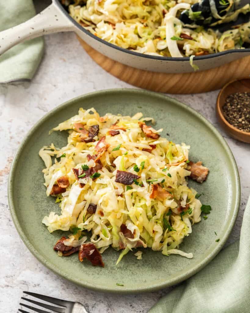 A single green plate full of fried cabbage with bacon and a skillet full of fried cabbage in the background.