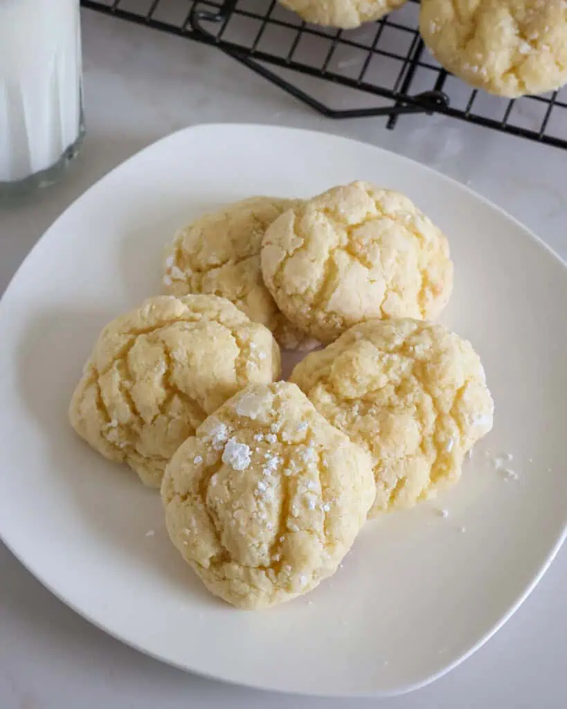 A serving plate full of gooey butter cookies with a glass of milk and the rest of the cookies in the background. 