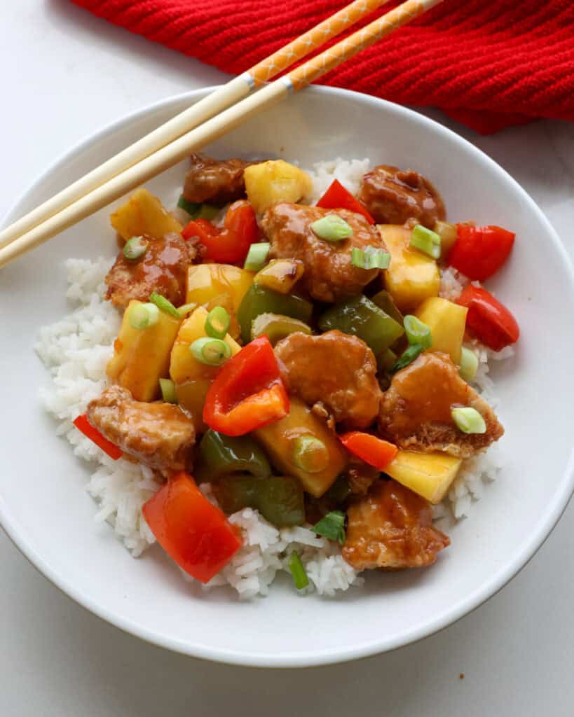 An overhead view of sweet and sour pork over rice on a plate with chopsticks in the background. 