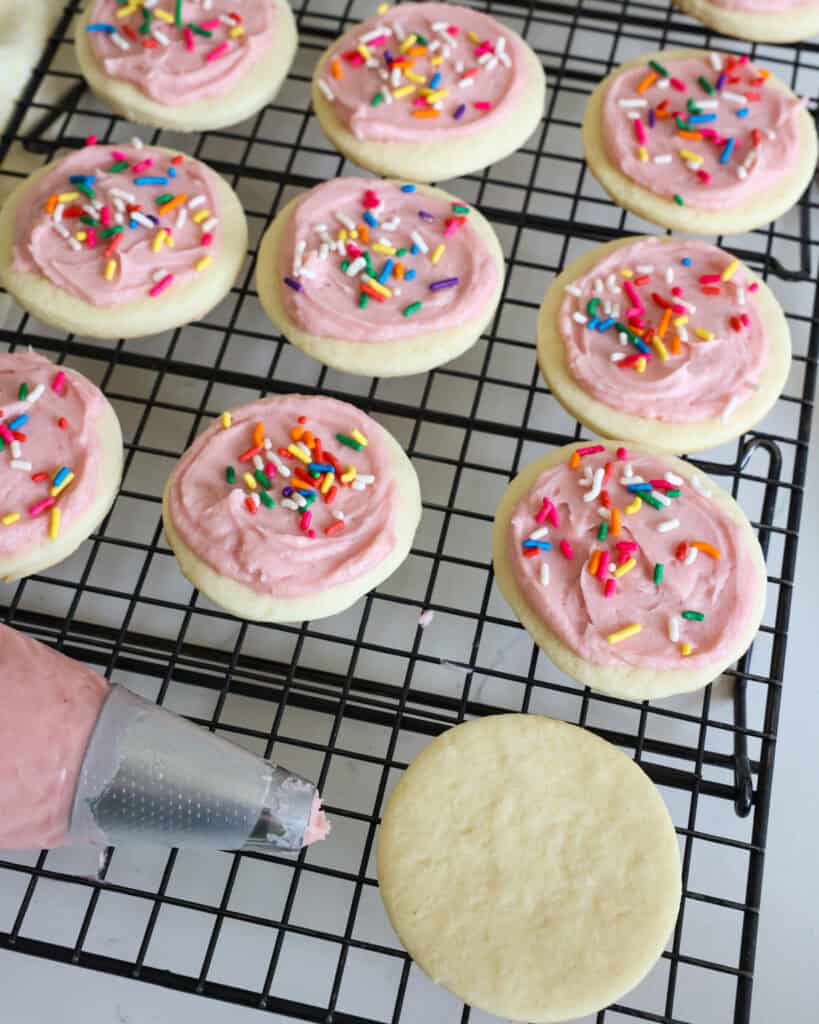 Lofthouse cookies on a wire rack with a pastry bag full of pink frosting and one cookie that is not iced. 